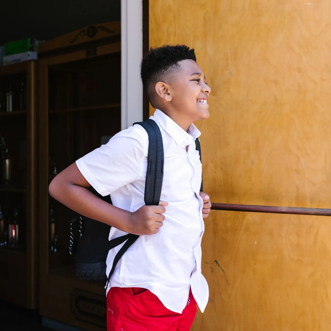 A smiling schoolboy in a white shirt with a black backpack stepping out through an open wooden school door in sunlight