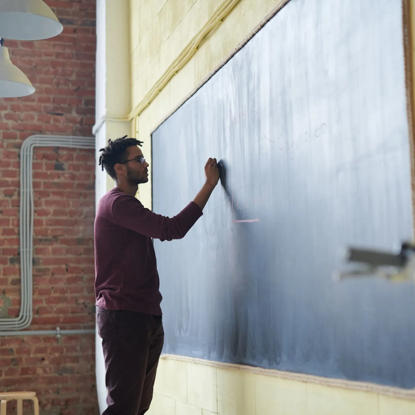 A teacher in a burgundy sweater writing on a large chalkboard in a sunlit classroom with brick and cream walls