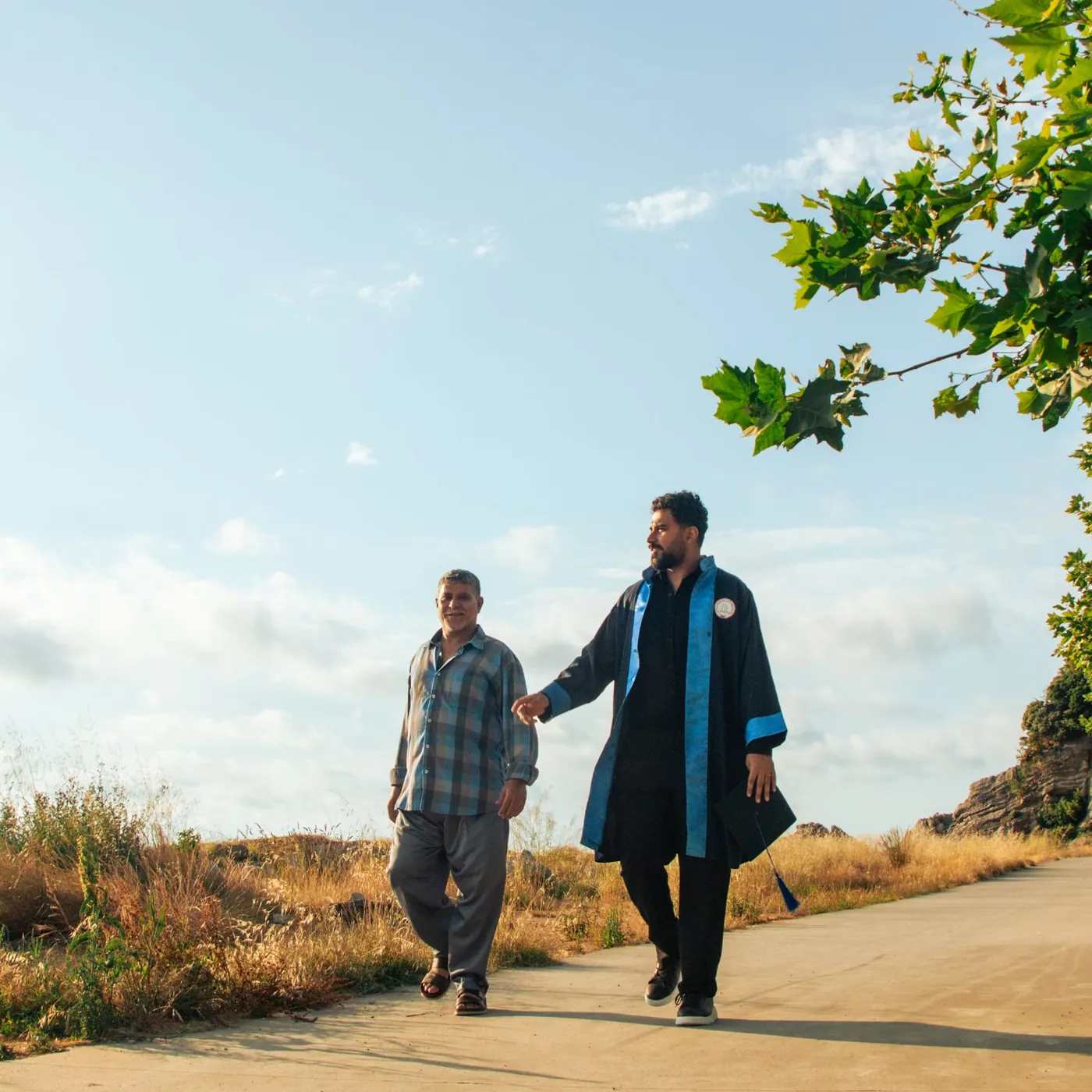 A young man in a graduation gown walking beside an older man along a sunny path lined with trees
