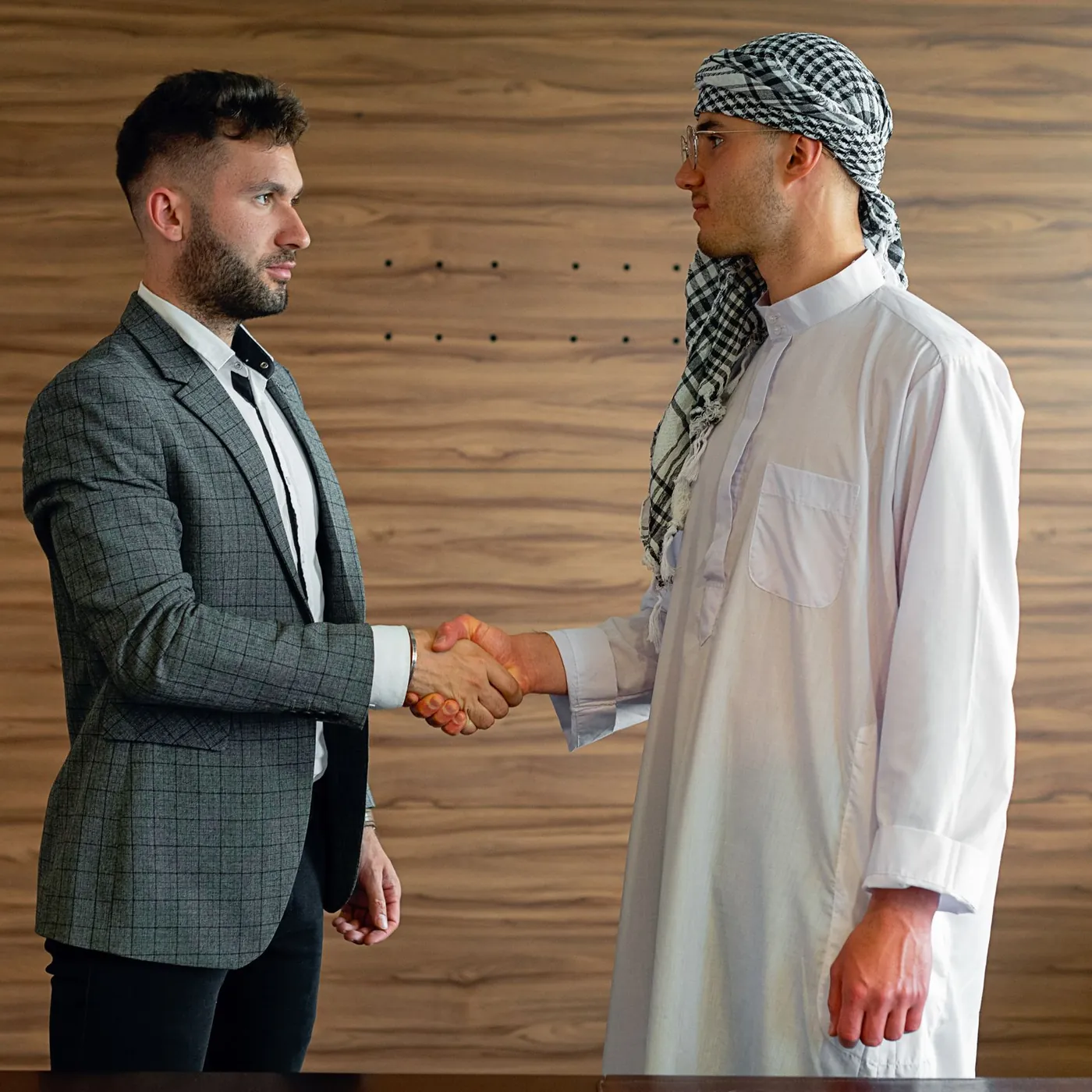 A man in a check blazer shaking hands with a man in a white thobe and keffiyeh against a warm wood-panelled wall