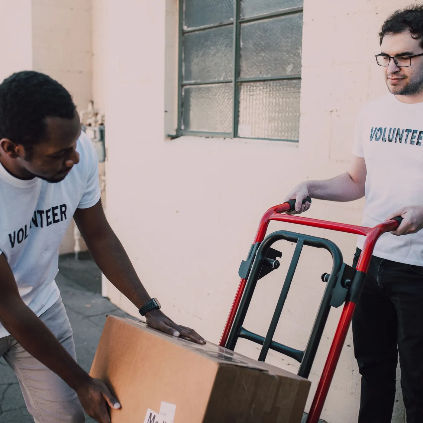 Two men in volunteer T-shirts lifting a cardboard box onto a red hand truck beside a cream-painted wall