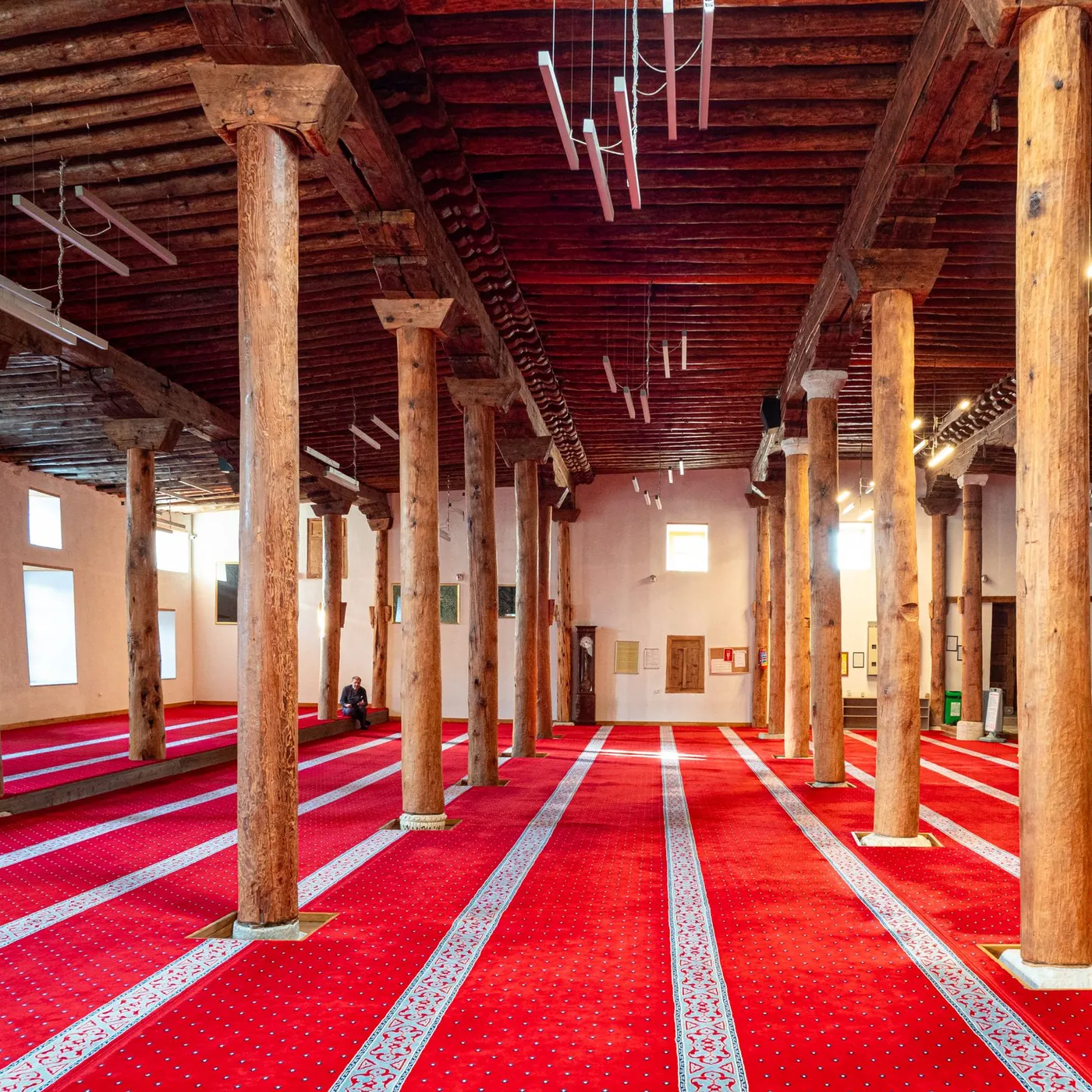 A wooden-pillared mosque hall with red prayer carpet in rows and a single man sitting quietly against the far wall