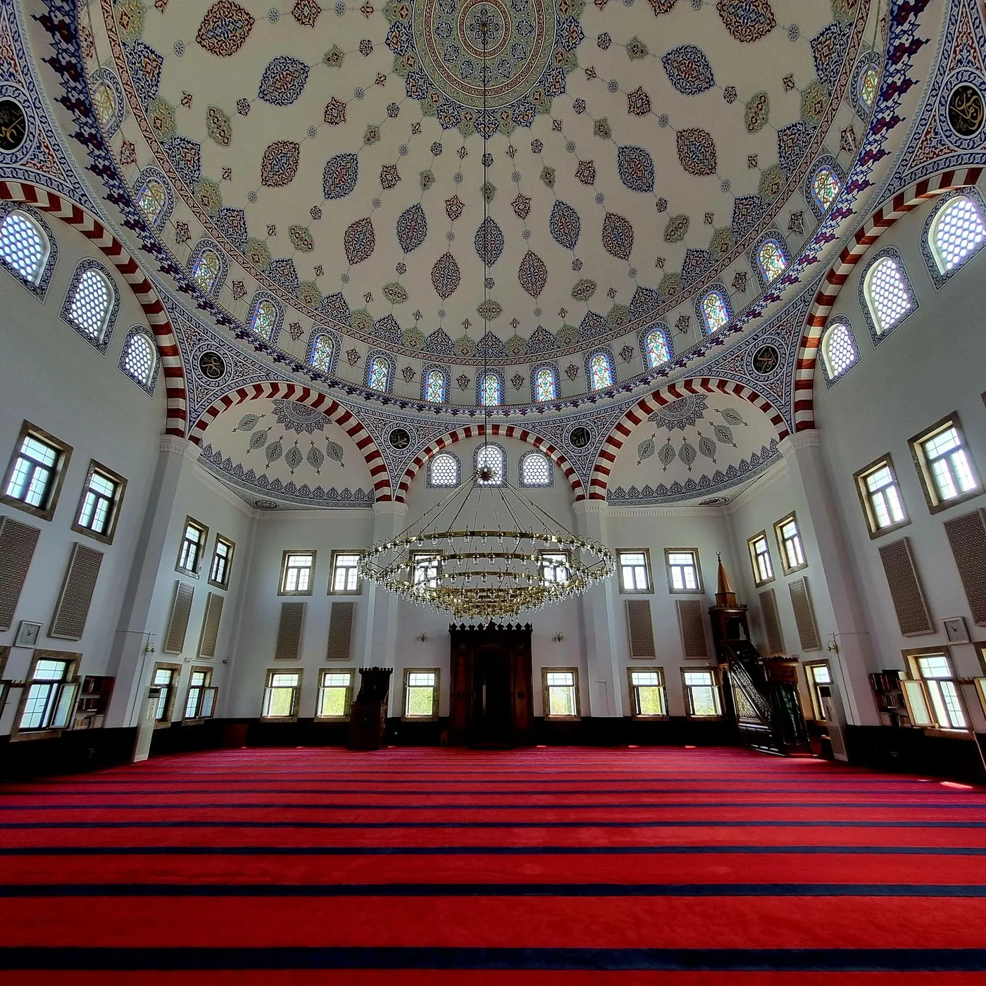 An empty mosque prayer hall under a painted dome, with red-striped carpet, a brass ring chandelier, mihrab and wooden minbar