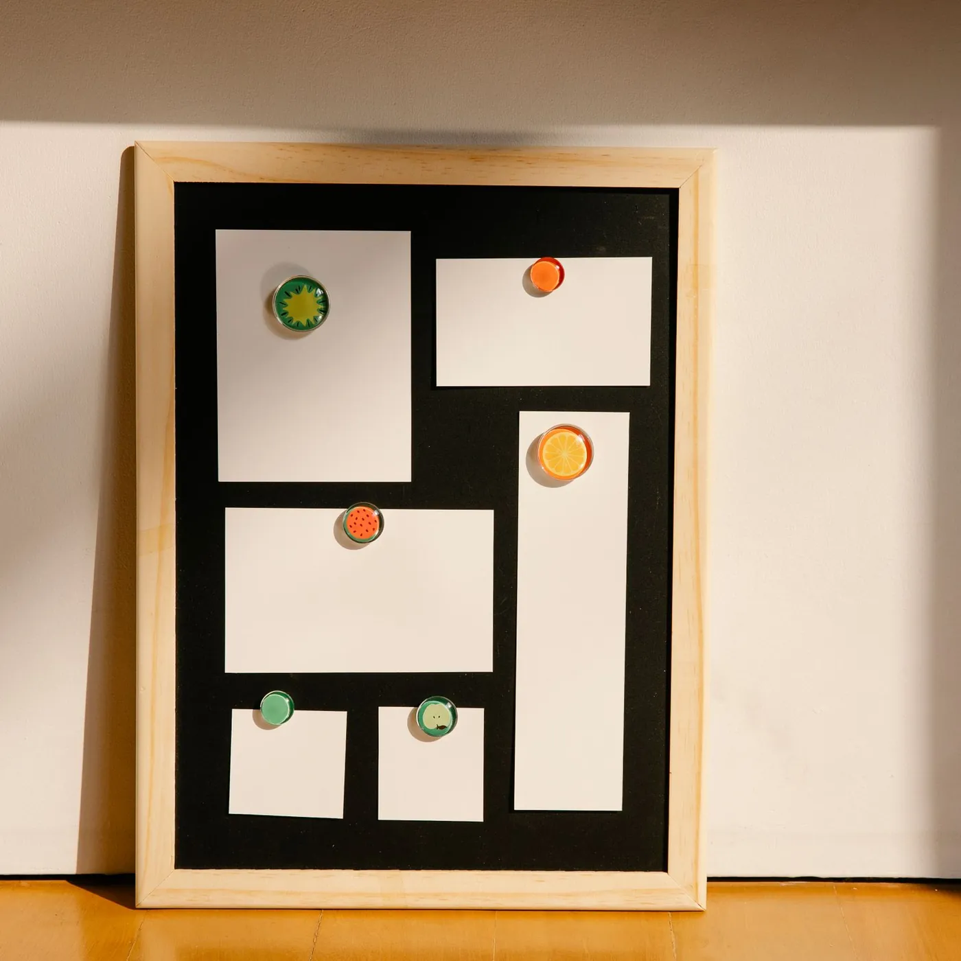 A pine-framed noticeboard with blank white cards held by fruit-shaped magnets, leaning against a sunlit wall