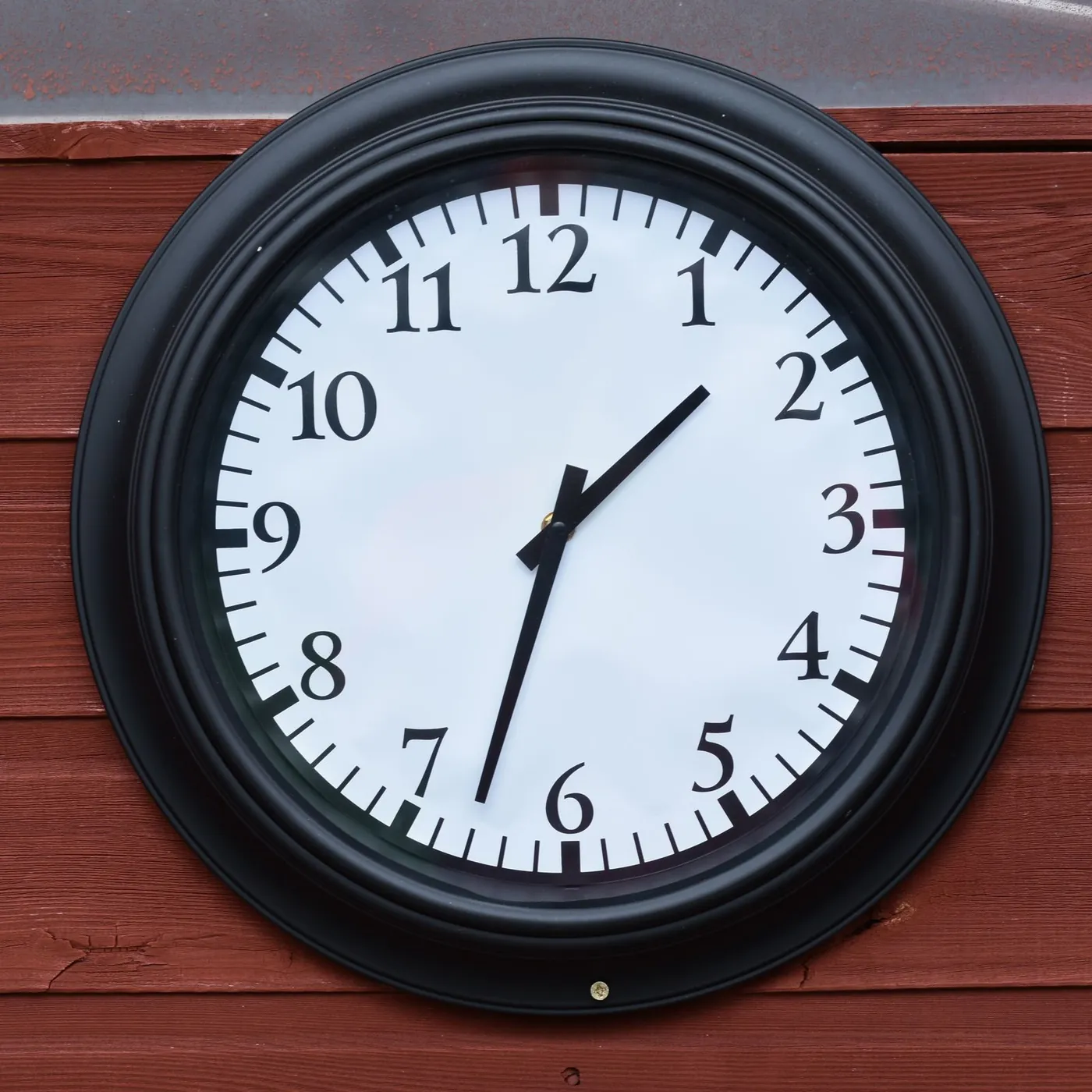 A plain black-framed wall clock with bold numerals mounted on red-painted wooden siding