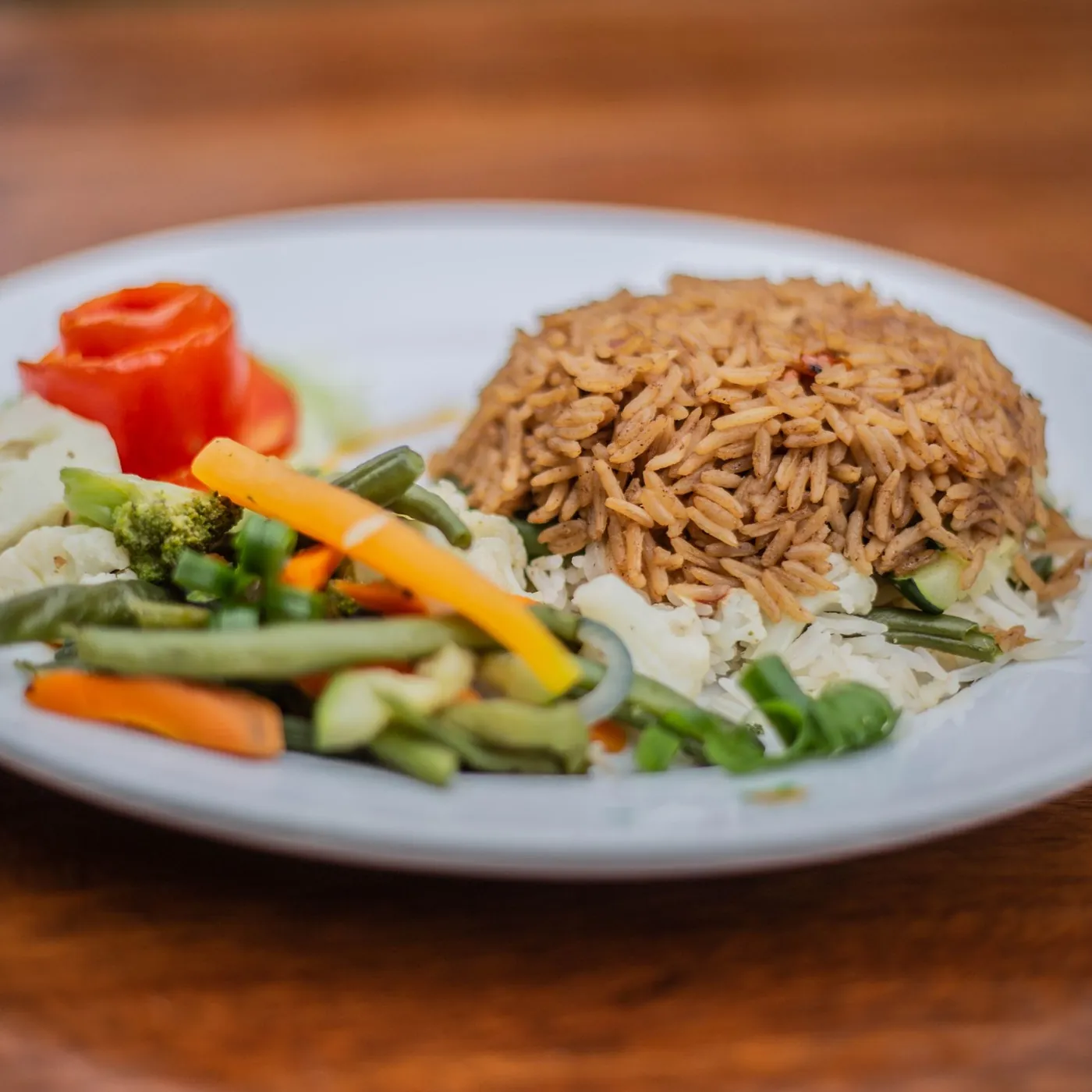 A plate of spiced rice with steamed green beans, carrot, cauliflower, broccoli and a tomato rose on a wooden table