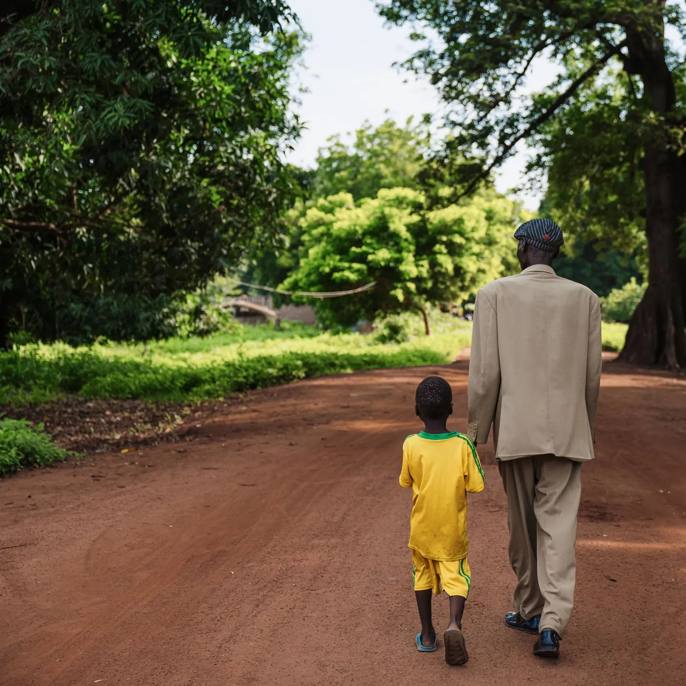 A father in a beige suit and cap walking hand in hand with his young son down a red dirt road under large trees