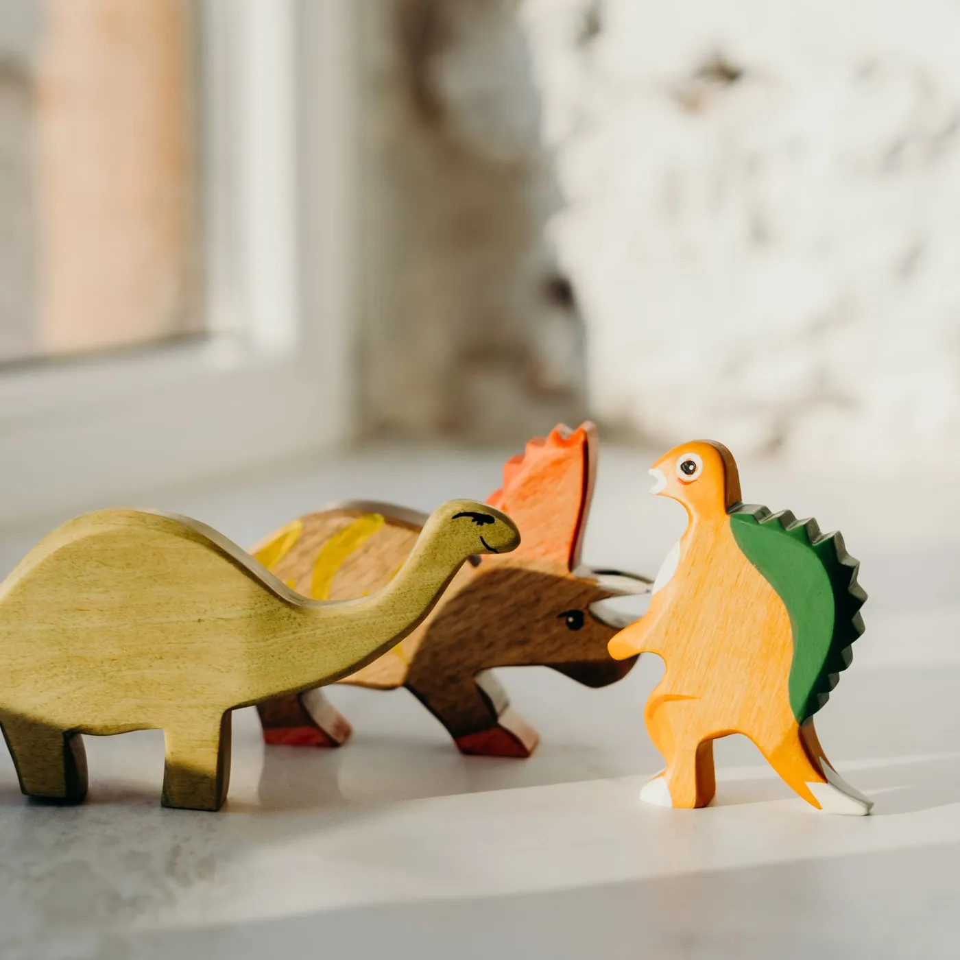 Three painted wooden toy dinosaurs, a long-necked green one, a triceratops and a small orange one, on a sunny windowsill