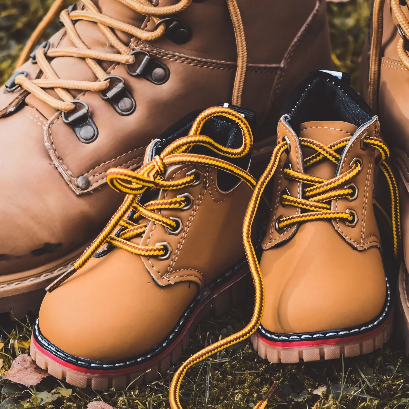 A small pair of tan lace-up children's boots standing between a pair of large, worn adult boots on autumn grass