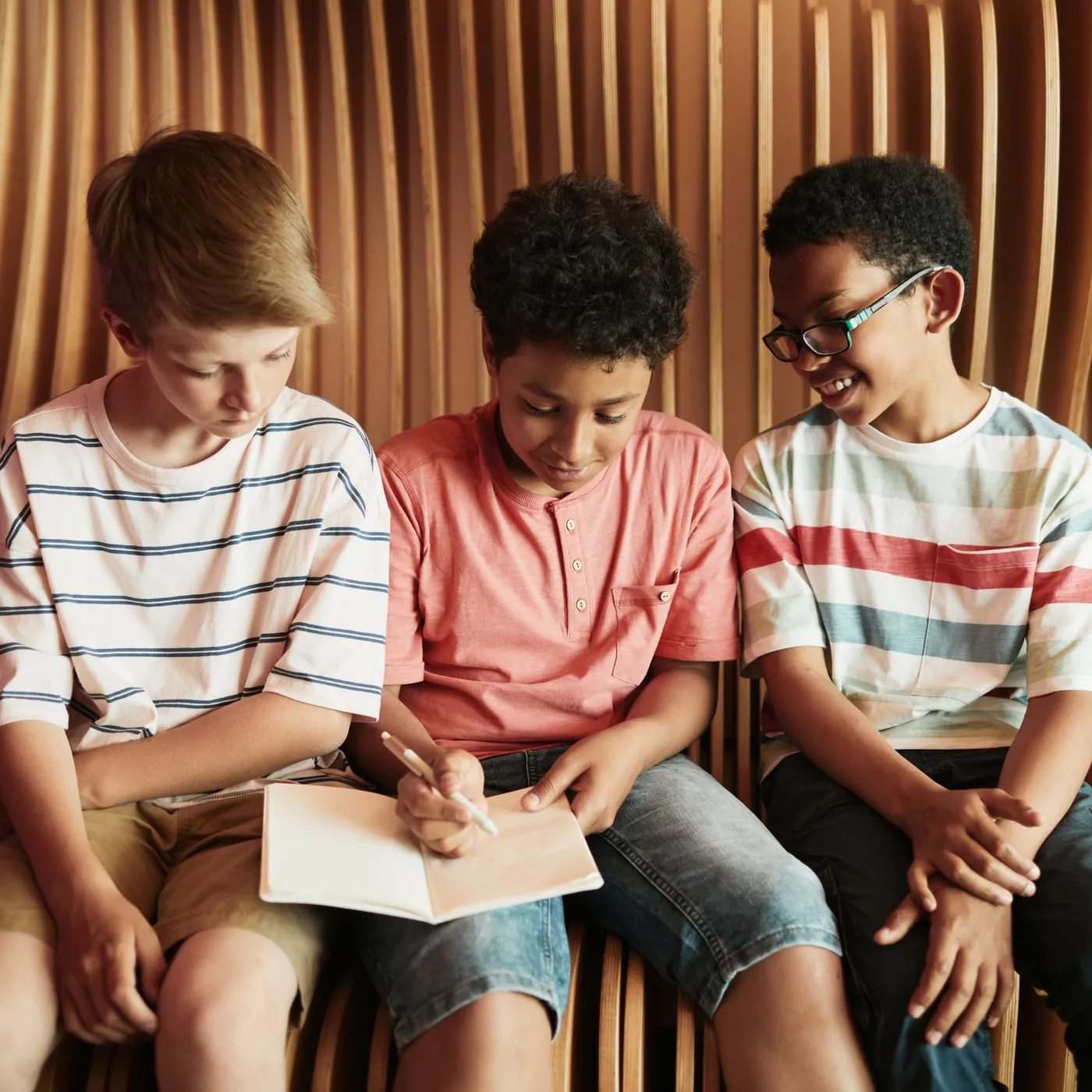 Three boys in striped T-shirts sitting close on a curved wooden bench, laughing as one writes in a notebook