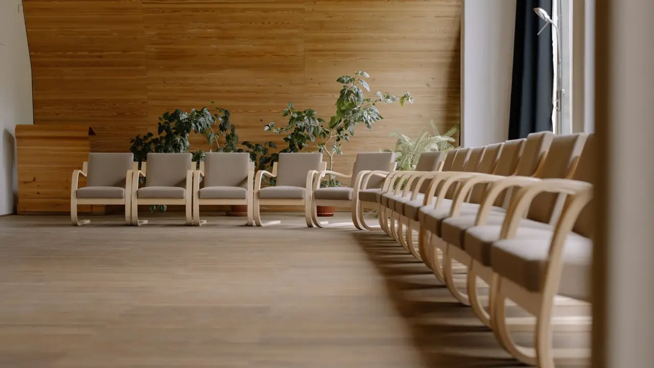 A warm pine-paneled hall with birch armchairs set out along the wall, living plants behind them and a long curving rank of matching chairs.