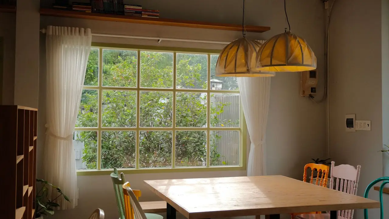 A plain scrubbed-pine table with mismatched painted chairs in sage, yellow, orange, cream and teal, under amber pendant lamps beside a tall window.