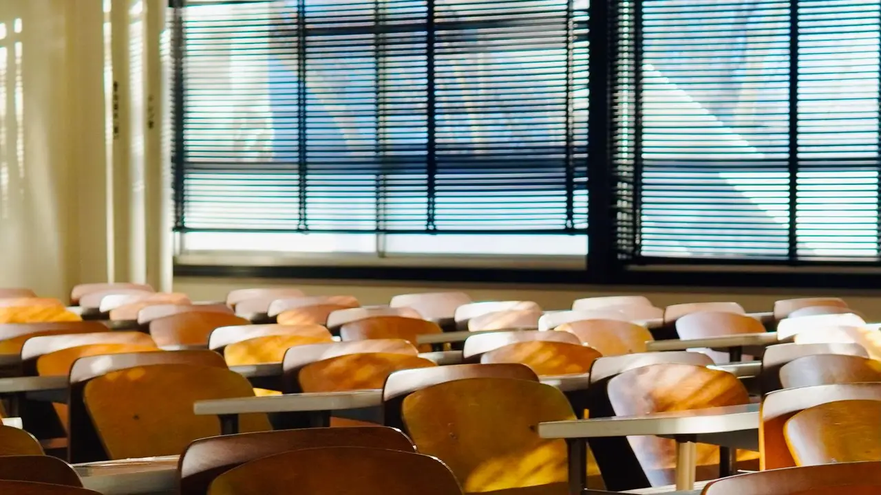 Rows of honey-oak tablet-arm school chairs in an empty classroom, late sun raking across the seat backs and venetian blinds behind.