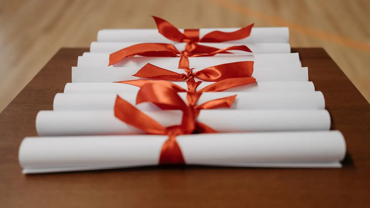 Eight documents rolled shut and tied with rust satin ribbon, ranked across a dark walnut table.