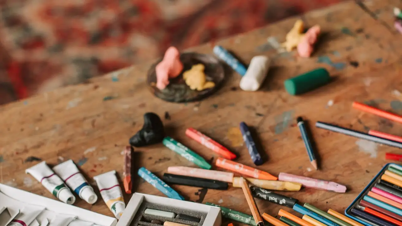 A well-used honey-wood craft table from above, with wax crayons, oil pastels, plasticine shapes and a tin of colored pencils.