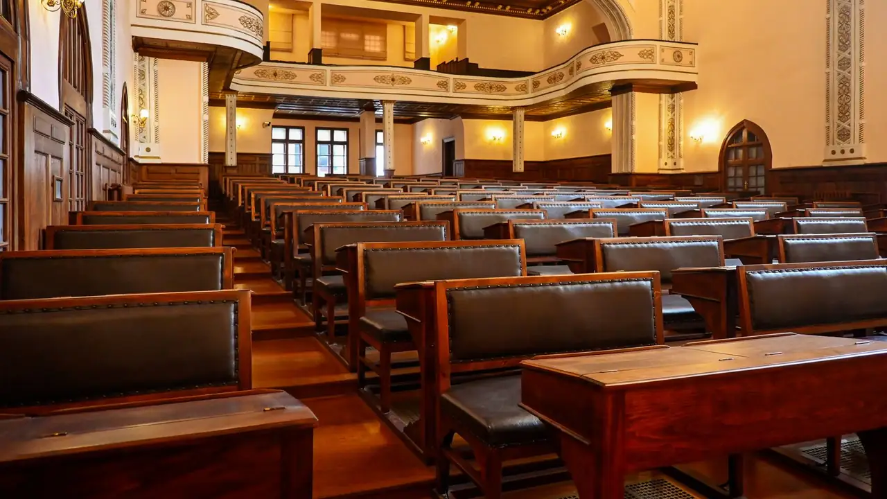 A historic assembly hall with mahogany bench desks and studded leather backs stepping up toward a balcony, brass sconces lit.