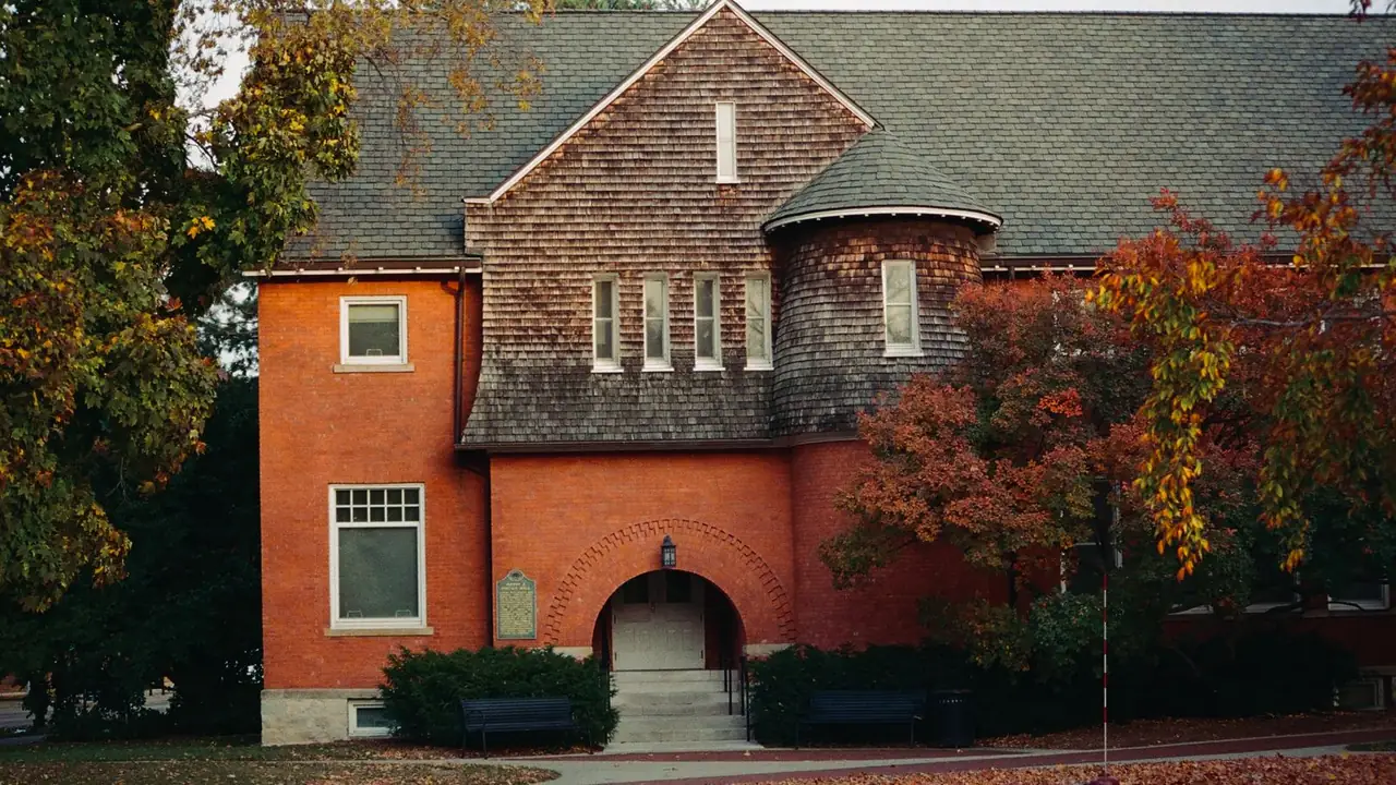 A red-brick and cedar-shingle campus building in autumn color, with an arched entrance and a shingled turret.