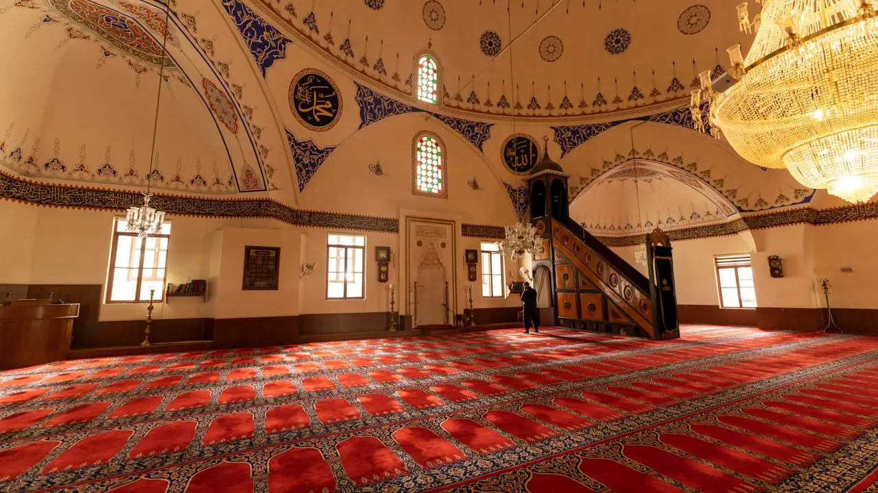 A bright prayer hall, the carpet marked into individual prayer spaces, with a carved wooden minbar and a crystal chandelier beyond.