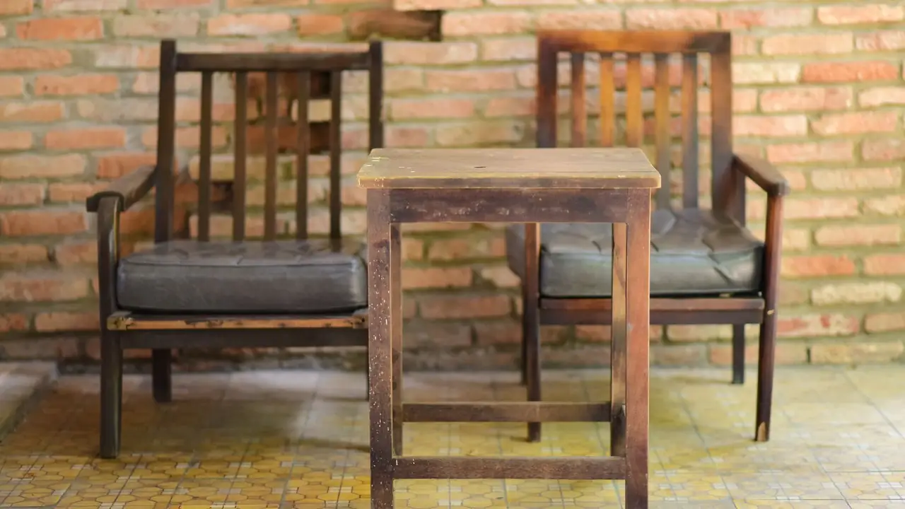 Two worn dark-wood armchairs either side of a small table, against a clean terracotta brick wall on ochre tile.