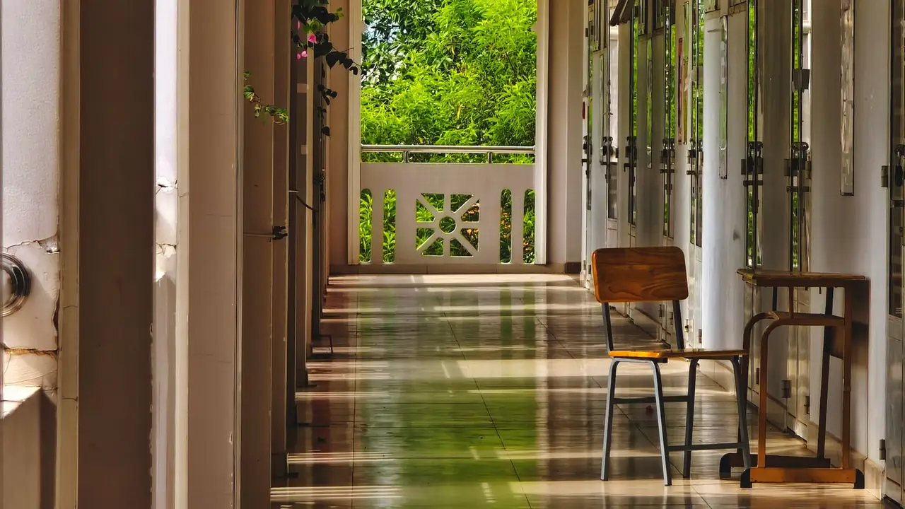 A sunlit school veranda with a wooden-seat school chair and a trestle desk against louvered classroom doors.