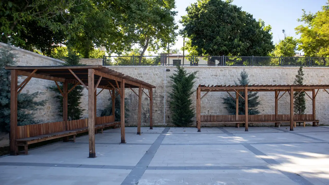 A walled sunlit school yard with timber pergolas over long runs of bench seating, cut stone and conifers behind.