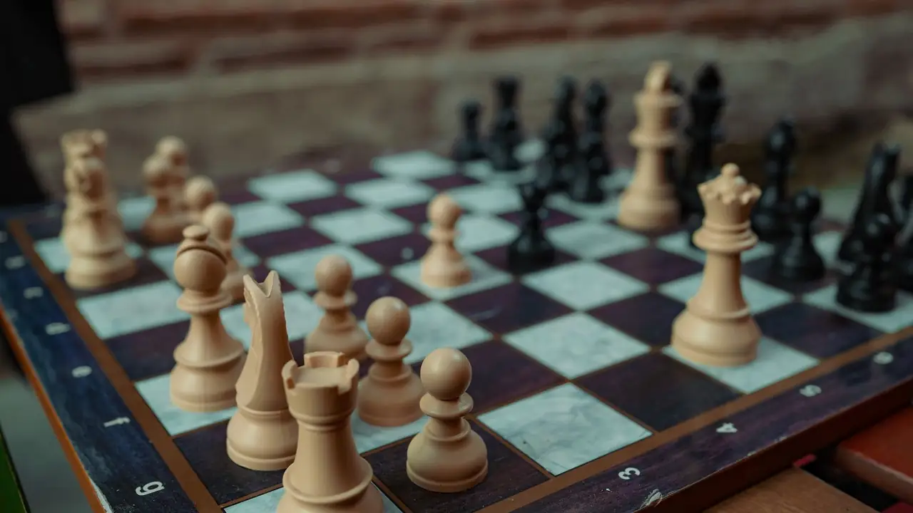A chess game in progress seen close, tan boxwood pieces on a walnut-edged board with warm brick blurred behind.