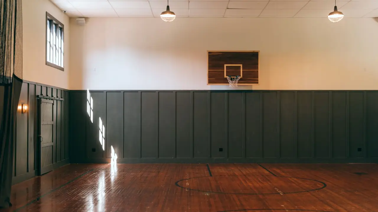An old school gymnasium with an oiled oak floor, olive board-and-batten dado, warm pendant lights and a timber-backed hoop.