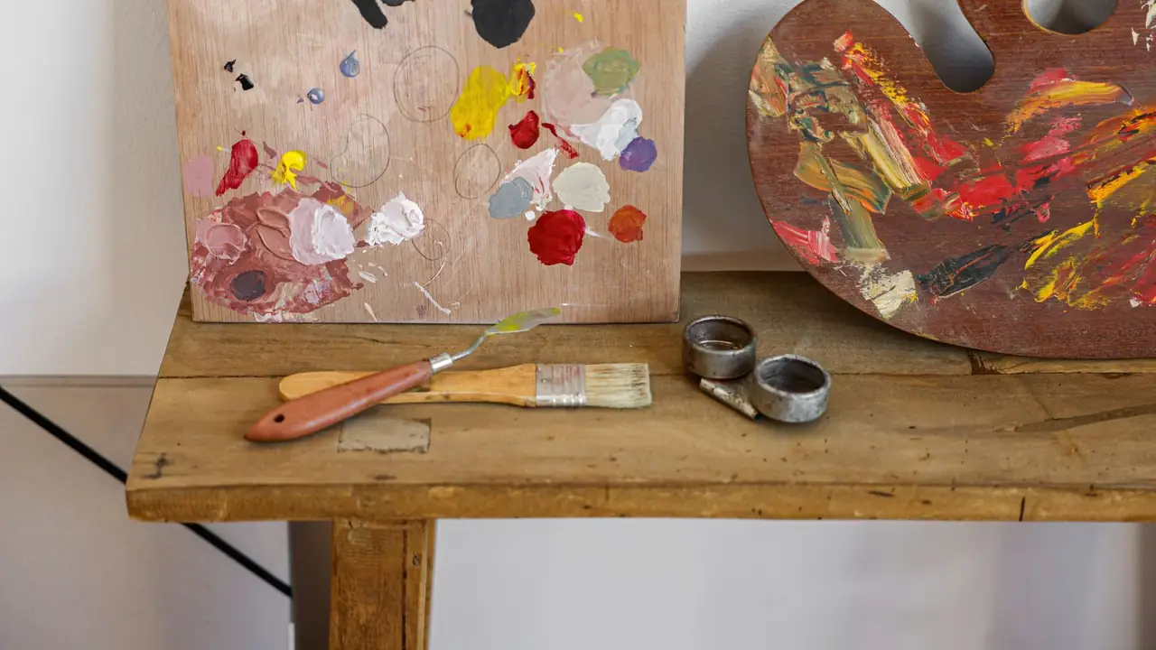 A rustic pine table with a paint-spattered mixing board, a palette knife and brush, and a loaded ochre and red palette.