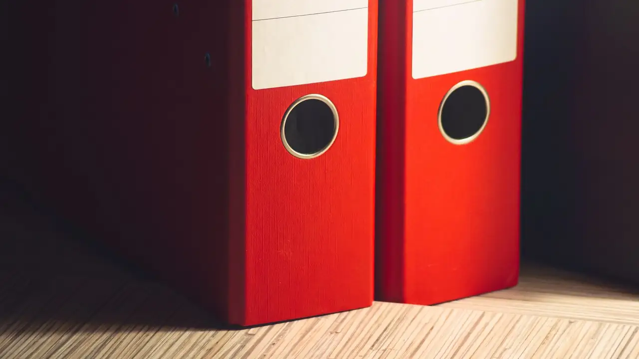 Two red lever-arch files standing upright on pale bamboo against a wall in deep shadow, raking light from the right.