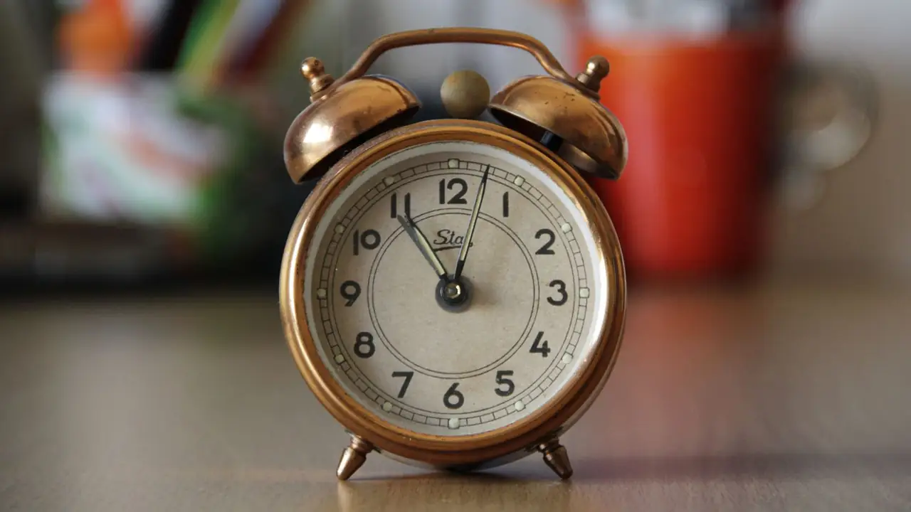A copper twin-bell alarm clock standing on a light wooden desk in warm daylight, a red mug blurred behind.