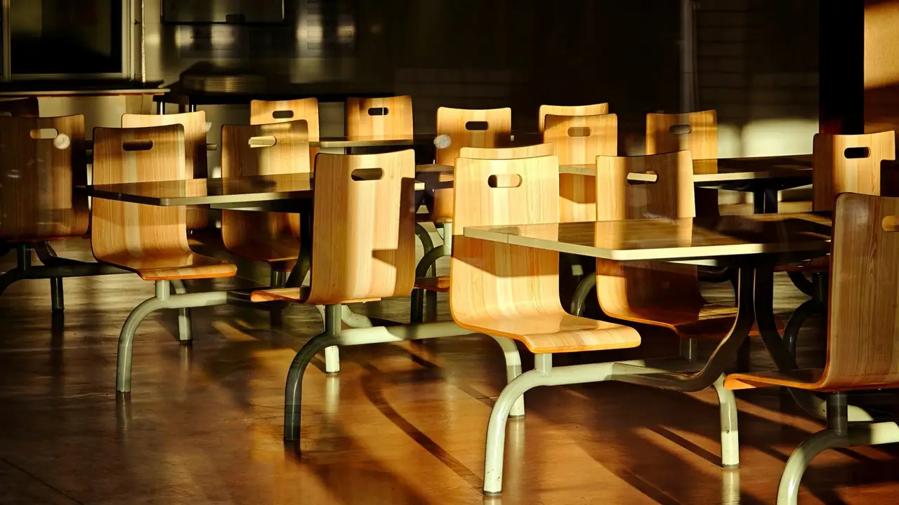 An empty school cafeteria in low golden sun, honey plywood bench-seat chairs casting long shadows across the floor.