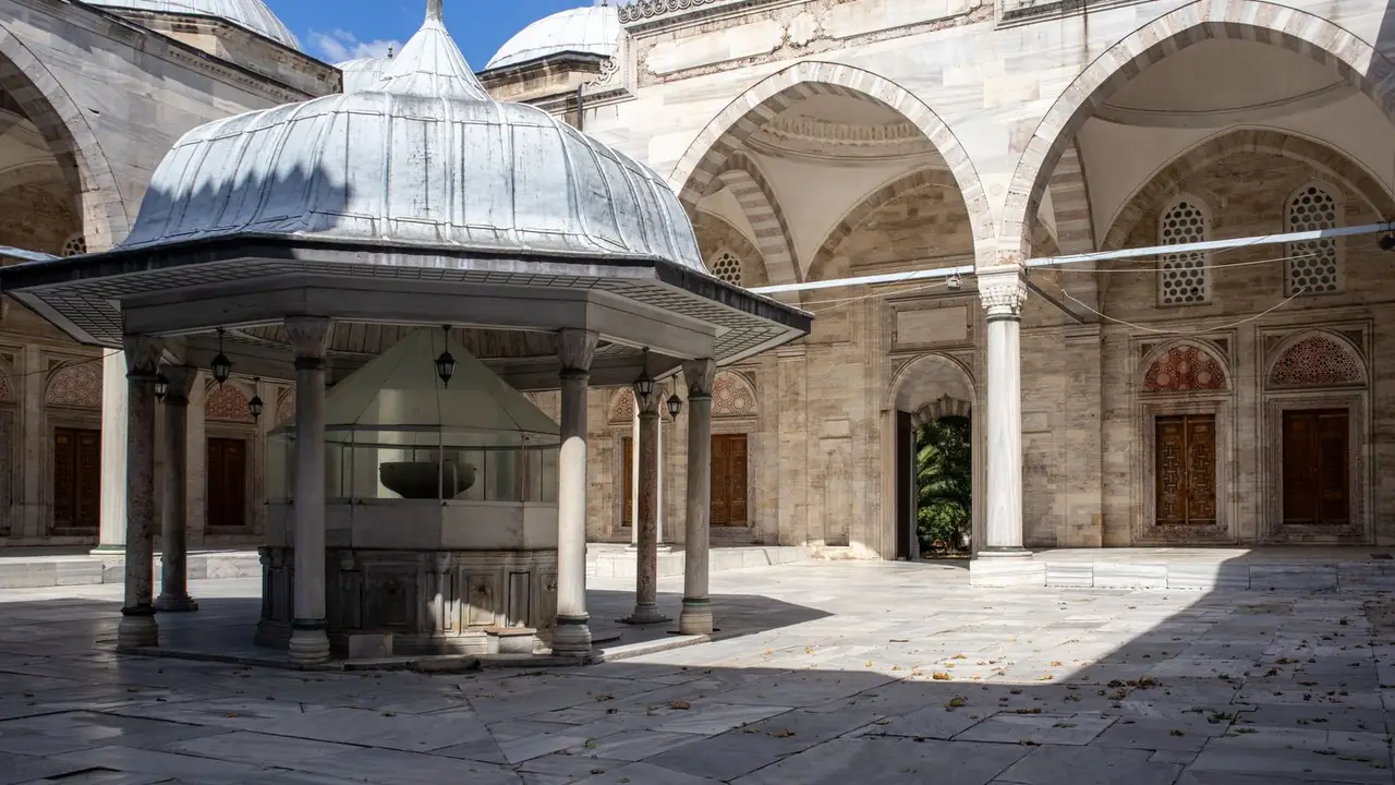 An Ottoman mosque courtyard with a domed ablution fountain at the center, a marble arcade and wooden doors beyond.