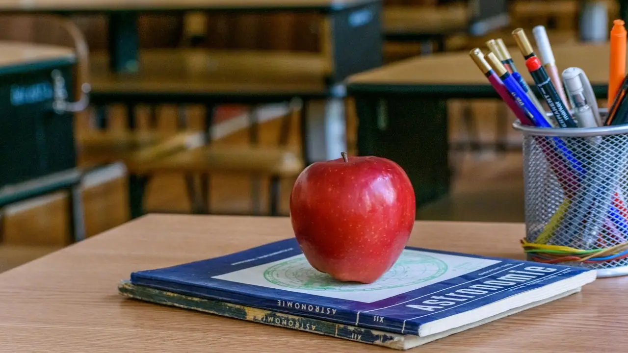 A red apple resting on a textbook on a pale oak classroom desk, a pot of pens beside it and desks soft in the background.